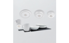 Wit servies op een donker rechthoekig onderlegger: twee mokken, twee kommen, gestapelde borden; drie ronde witte schijven bovenaan