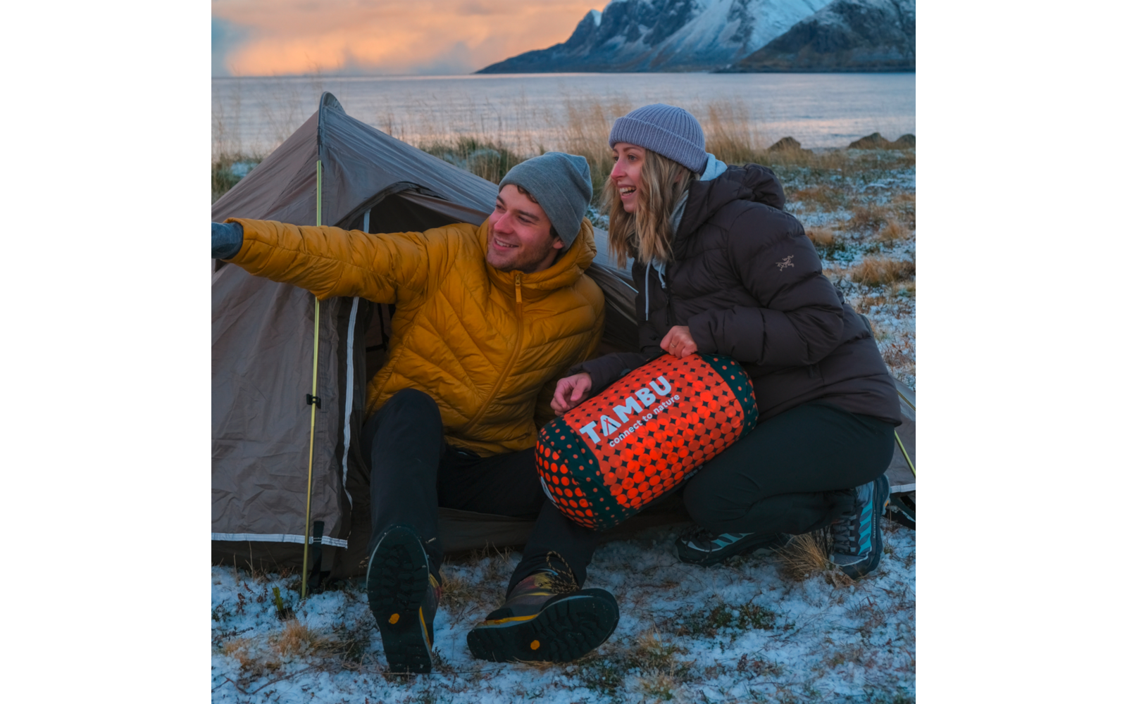Two people sit in front of a small tent on snowy ground by a shoreline, with water and mountains in the background; one person points with an outstretched arm, the other holds an orange-black sleeping bag labeled TAMBU. They wear winter jackets and beanies