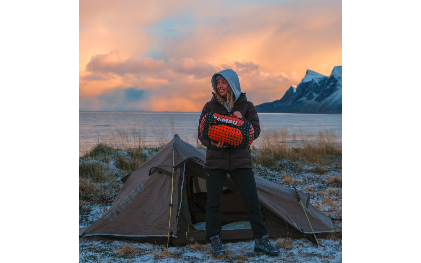 Person wearing a hood stands in front of a pitched tent holding an orange-and-black rolled sleeping bag labeled 'AMBU'; background shows sea, mountains, frost-covered grass and an orange sky
