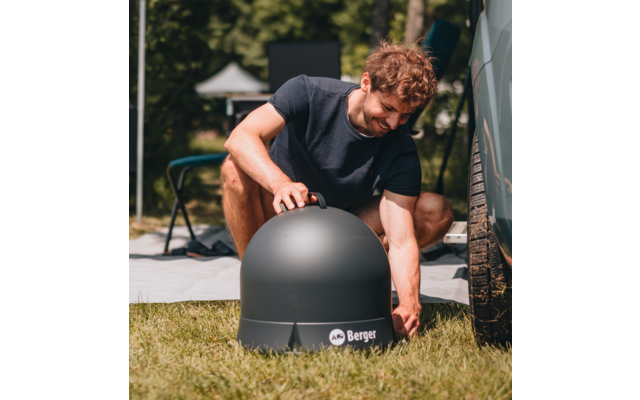 Bearded man in black t-shirt kneeling on grass setting up a black dome near a vehicle