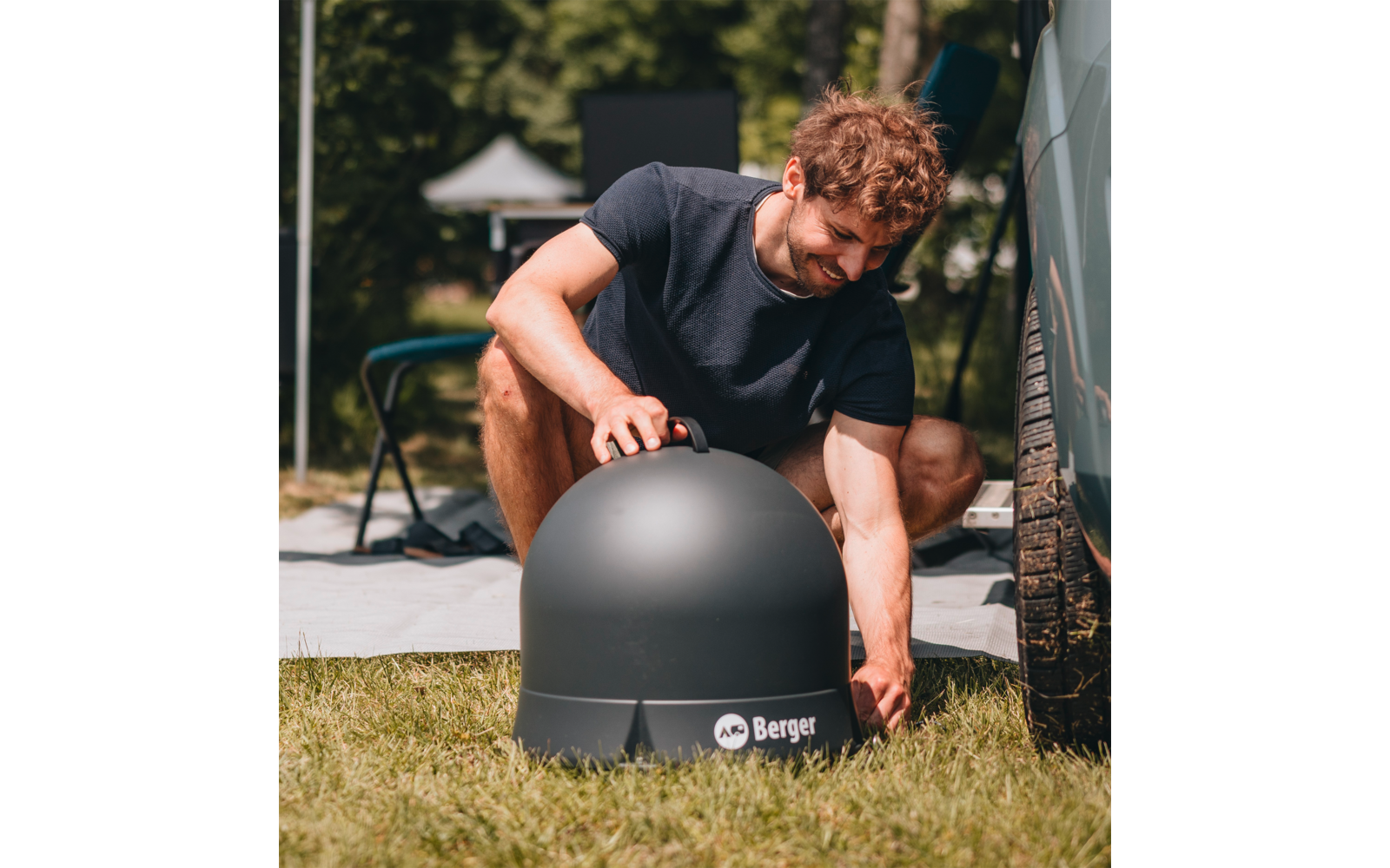 Bearded man in black t-shirt kneeling on grass setting up a black dome near a vehicle