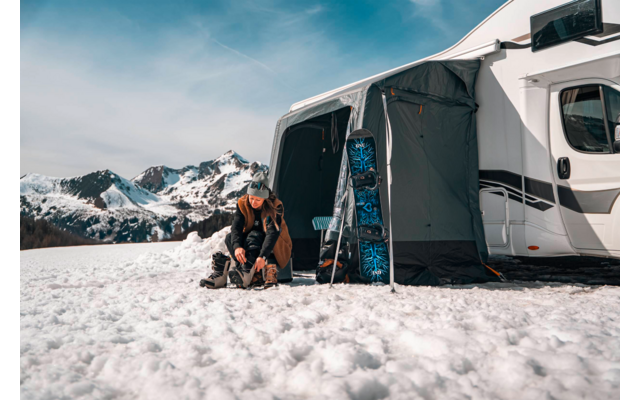 Camper met aangebouwde luifel op sneeuw, een persoon knielt en trekt laarzen aan, een snowboard leunt tegen de luifel, besneeuwde bergen en blauwe lucht op de achtergrond