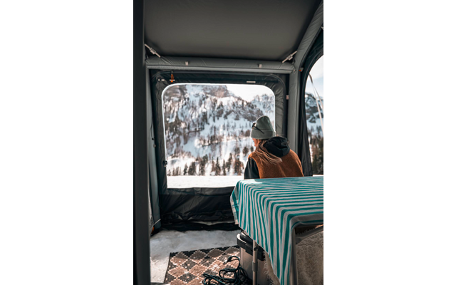 Persoon van achteren met muts en bruine jas zit aan een tafel in een binnenruimte; door een grote raamopening zijn besneeuwde bergen en bomen zichtbaar; tafel met gestreept kleed