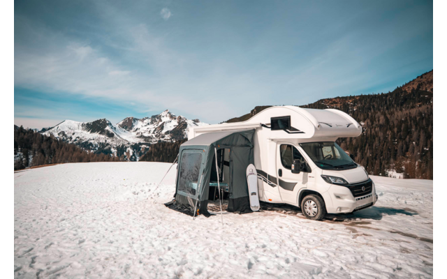 Camper bianco con tenda annessa su un campo coperto di neve, bosco di conifere e montagne innevate sullo sfondo sotto un cielo azzurro