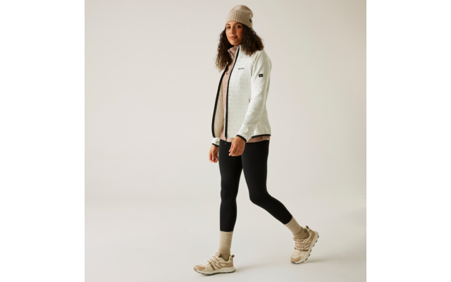 Frau in weißer Jacke, schwarzer Hose, beige Mütze und hellen Schuhen vor weißem Hintergrund