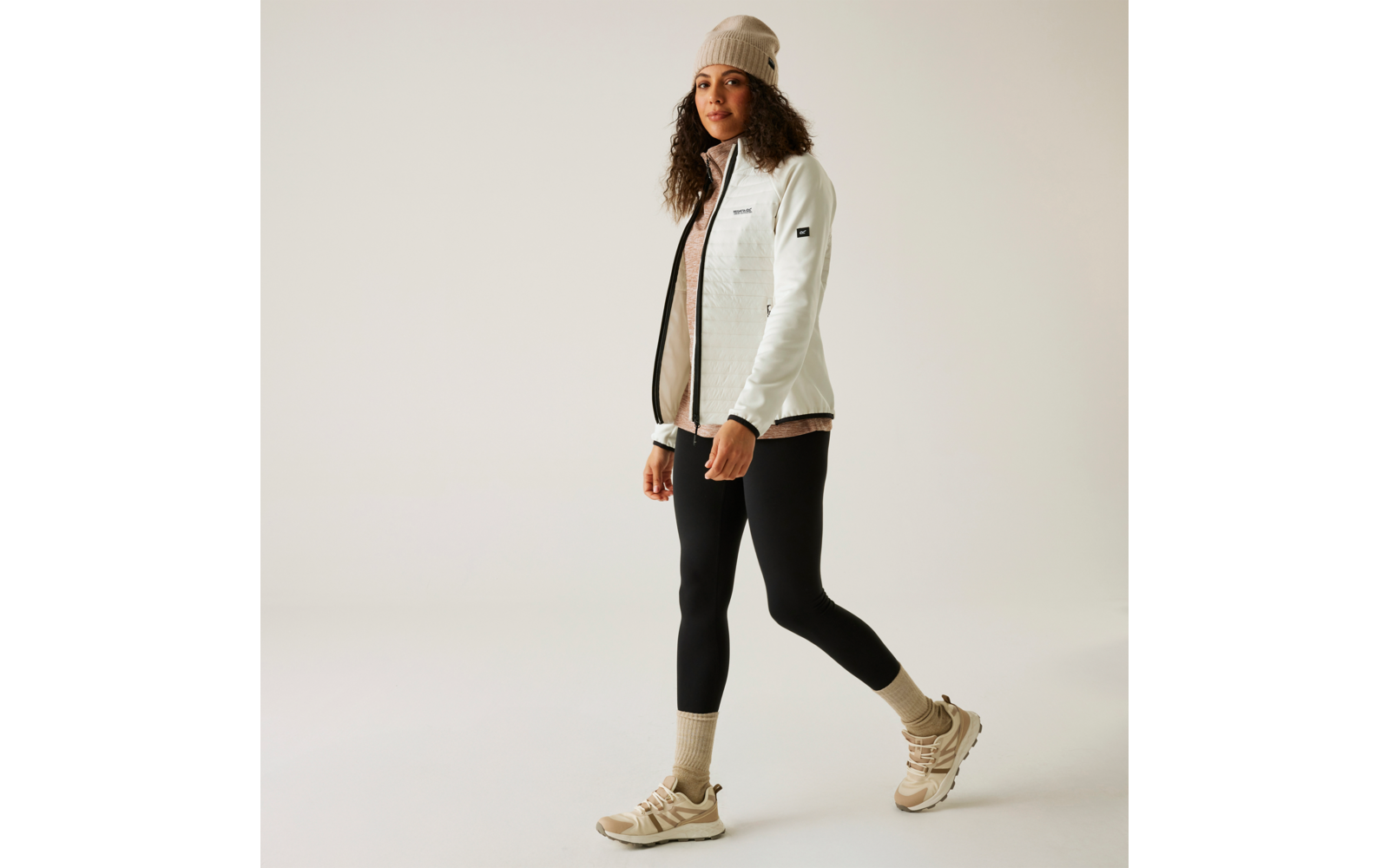 Frau in weißer Jacke, schwarzer Hose, beige Mütze und hellen Schuhen vor weißem Hintergrund