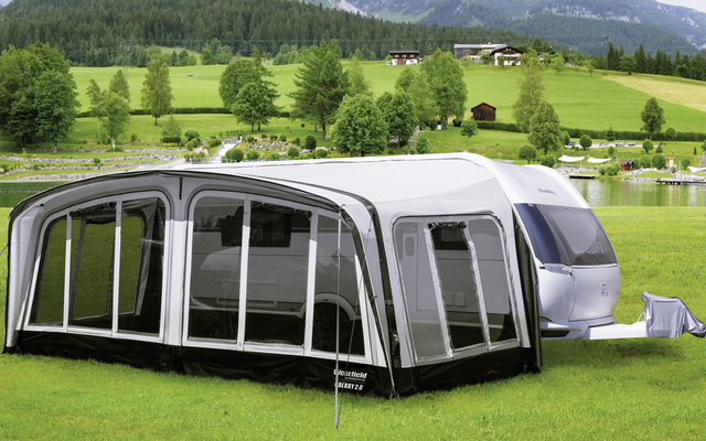 Caravana con toldo desmontable en un prado verde frente a colinas boscosas y montañas