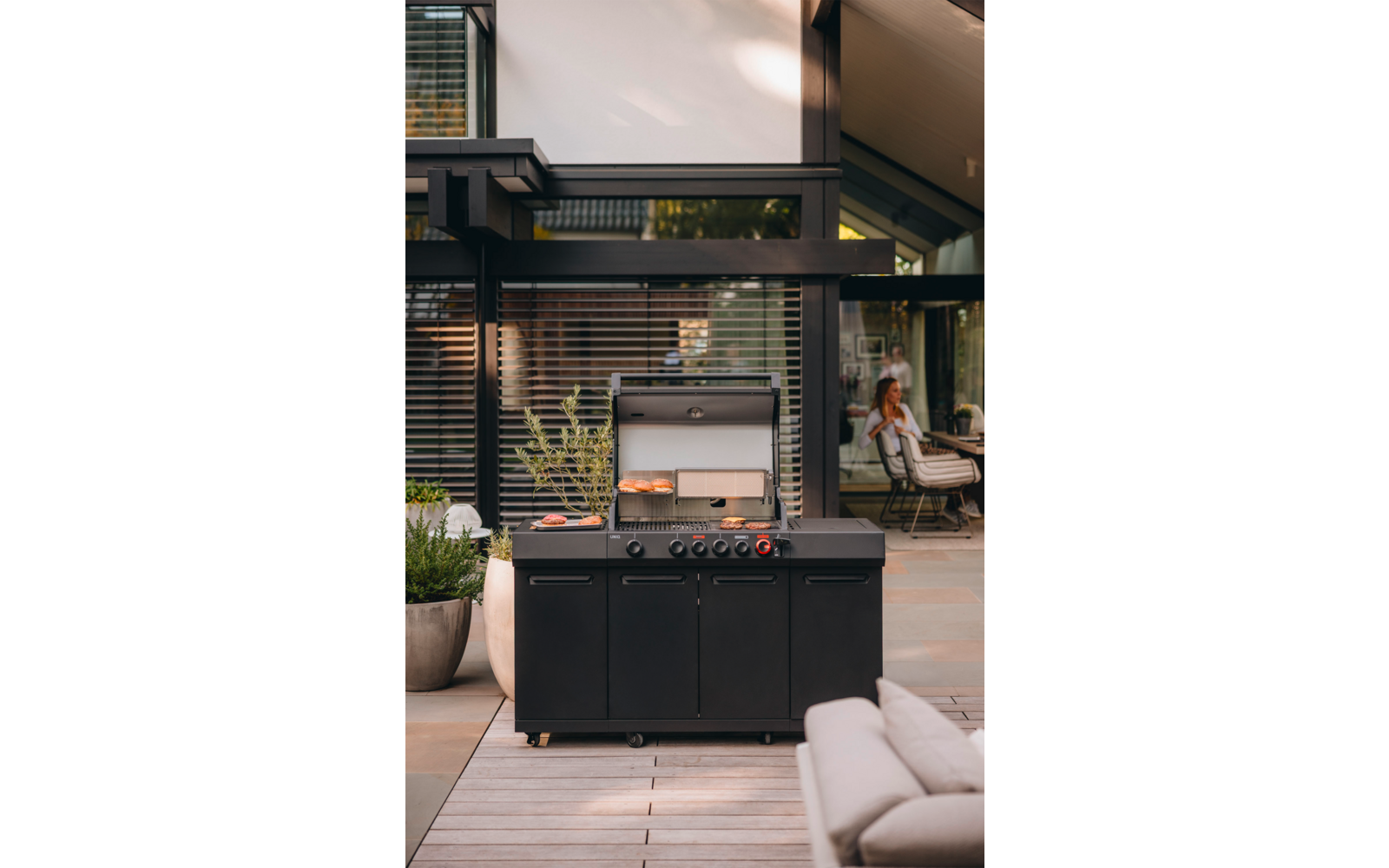 Schwarzer Gasgrill mit offenem Deckel auf Terrasse, zwei Brötchen auf dem Rost, Pflanztöpfe links, modernes Haus mit Glasfassade und eine Person sitzt im Hintergrund innen