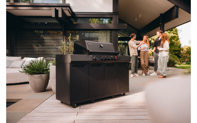 Schwarzer Gasgrill auf Holzterrasse im Vordergrund; links Pflanzen und Lounge-Sofa; im Hintergrund rechts drei Personen vor einem Haus mit horizontalen Fensterlamellen