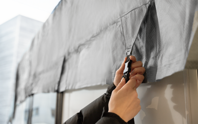Two hands hold a black utility knife cutting an opening in a gray fabric or tarp in front of a glass window; blurred buildings in the background
