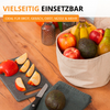 Panier en tissu beige contenant bananes, pommes, avocat, poire et nectarine sur une table en bois; à côté, quartiers de pomme sur une planche foncée, un couteau, un avocat sur un tissu et deux bocaux en verre en arrière‑plan
