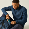 Sitzender Mann steckt eine Trinkflasche in einen blauen Rucksack; trägt einen dunkelblauen Fleecepullover mit kurzem Reißverschluss und dunkle Hose vor hellem Hintergrund