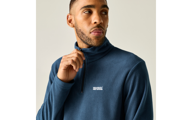 Oberkörper eines Mannes in blauem Fleece mit halbem Reißverschluss, Hand hält den Reißverschluss, er blickt zur Seite, heller Hintergrund