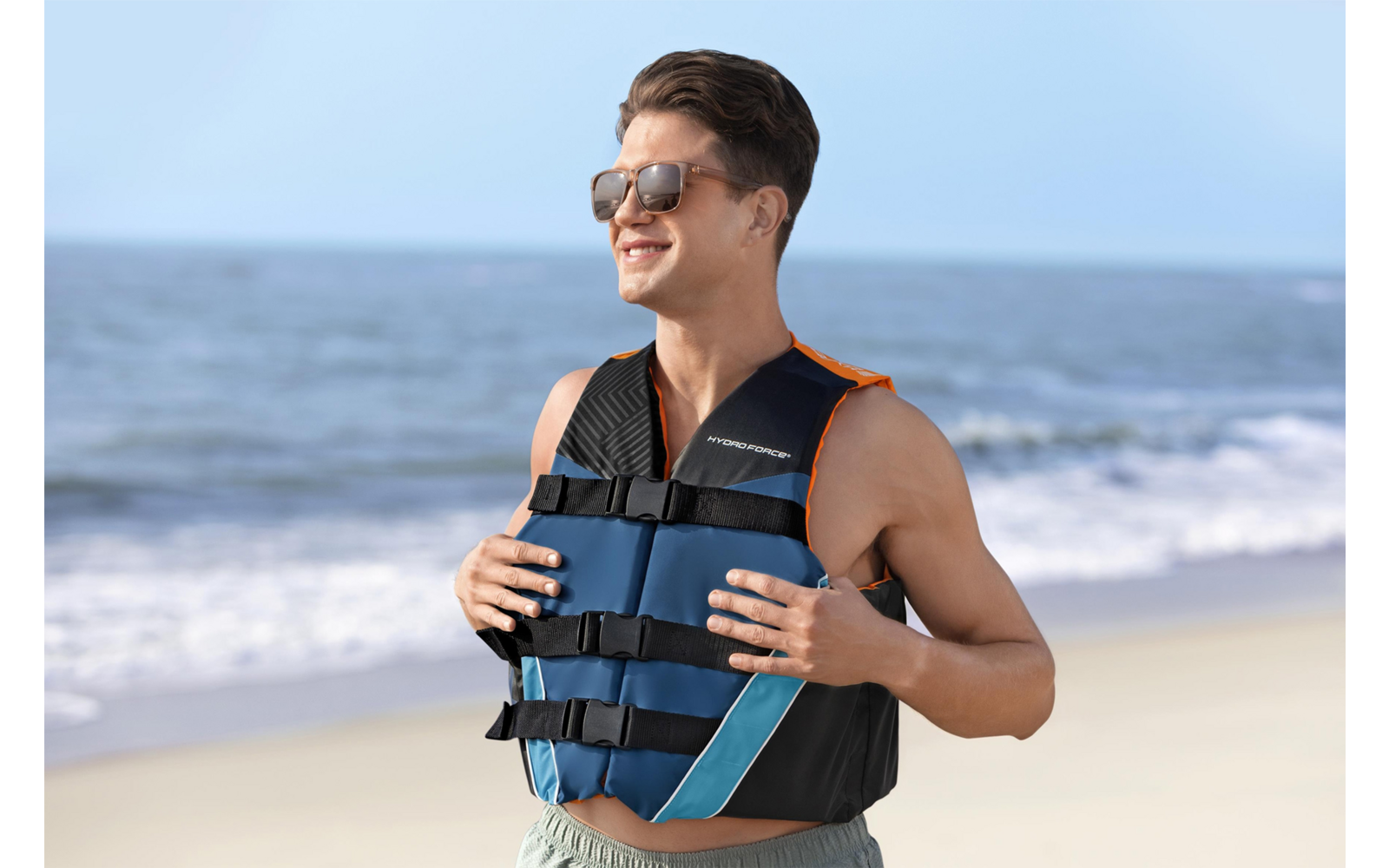 Person mit Schwimmweste und Sonnenbrille steht auf einem Sandstrand und hält die Westenriemen; Meer und Himmel im Hintergrund