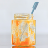 Bocal en verre contenant une pâte orange brillante avec des traces sur les parois intérieures; cuillère à manche bleu à l'intérieur; posé sur une surface réfléchissante devant un fond clair