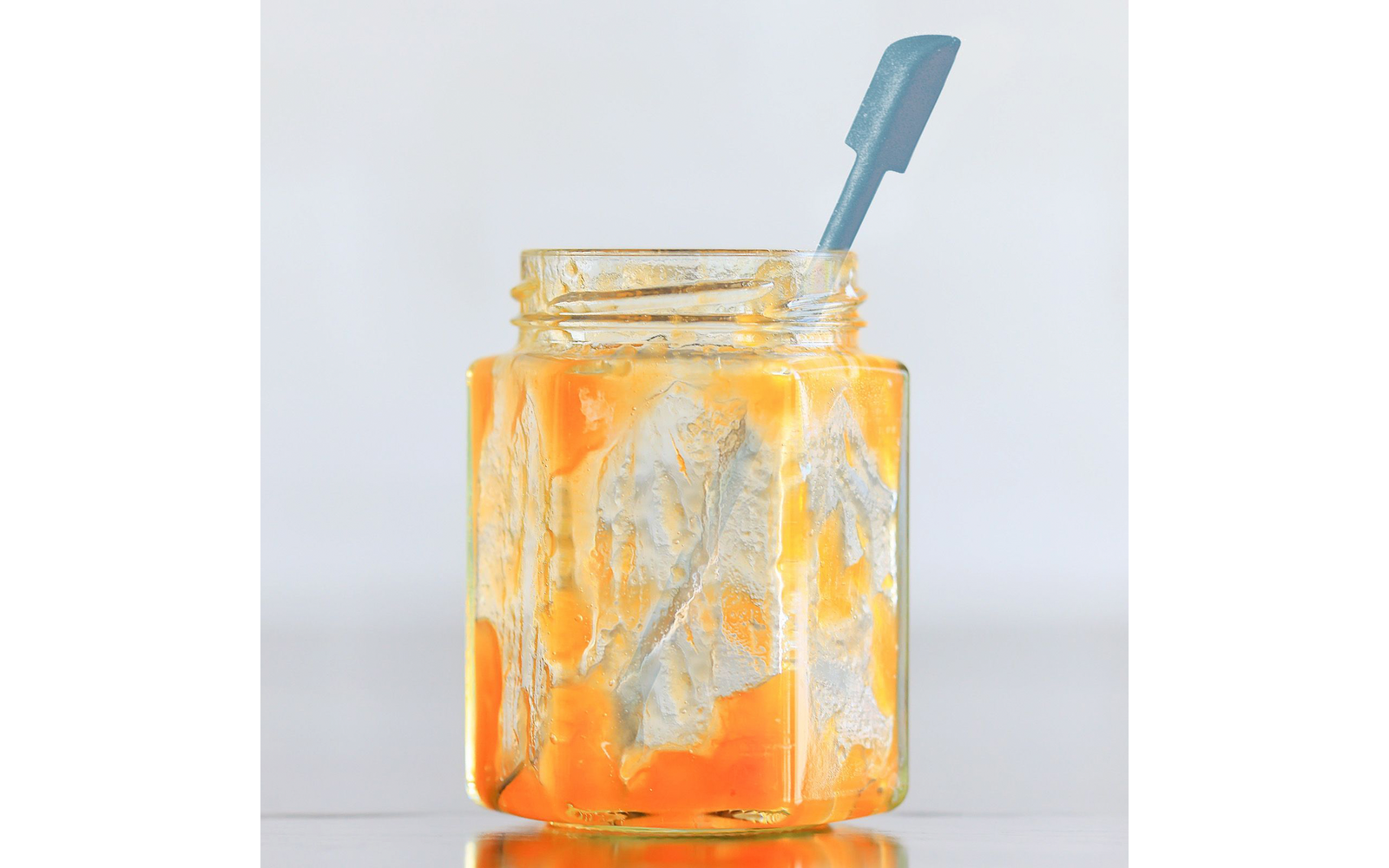 Bocal en verre contenant une pâte orange brillante avec des traces sur les parois intérieures; cuillère à manche bleu à l'intérieur; posé sur une surface réfléchissante devant un fond clair