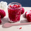 Bocal ouvert de confiture de framboise rouge sur un support clair, avec des framboises et une cuillère rose à côté