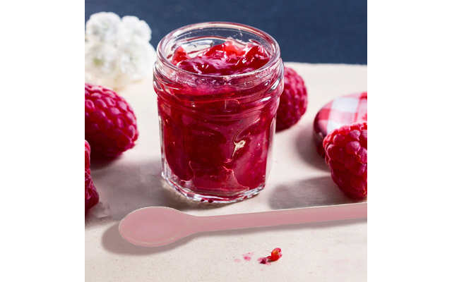 Bocal ouvert de confiture de framboise rouge sur un support clair, avec des framboises et une cuillère rose à côté