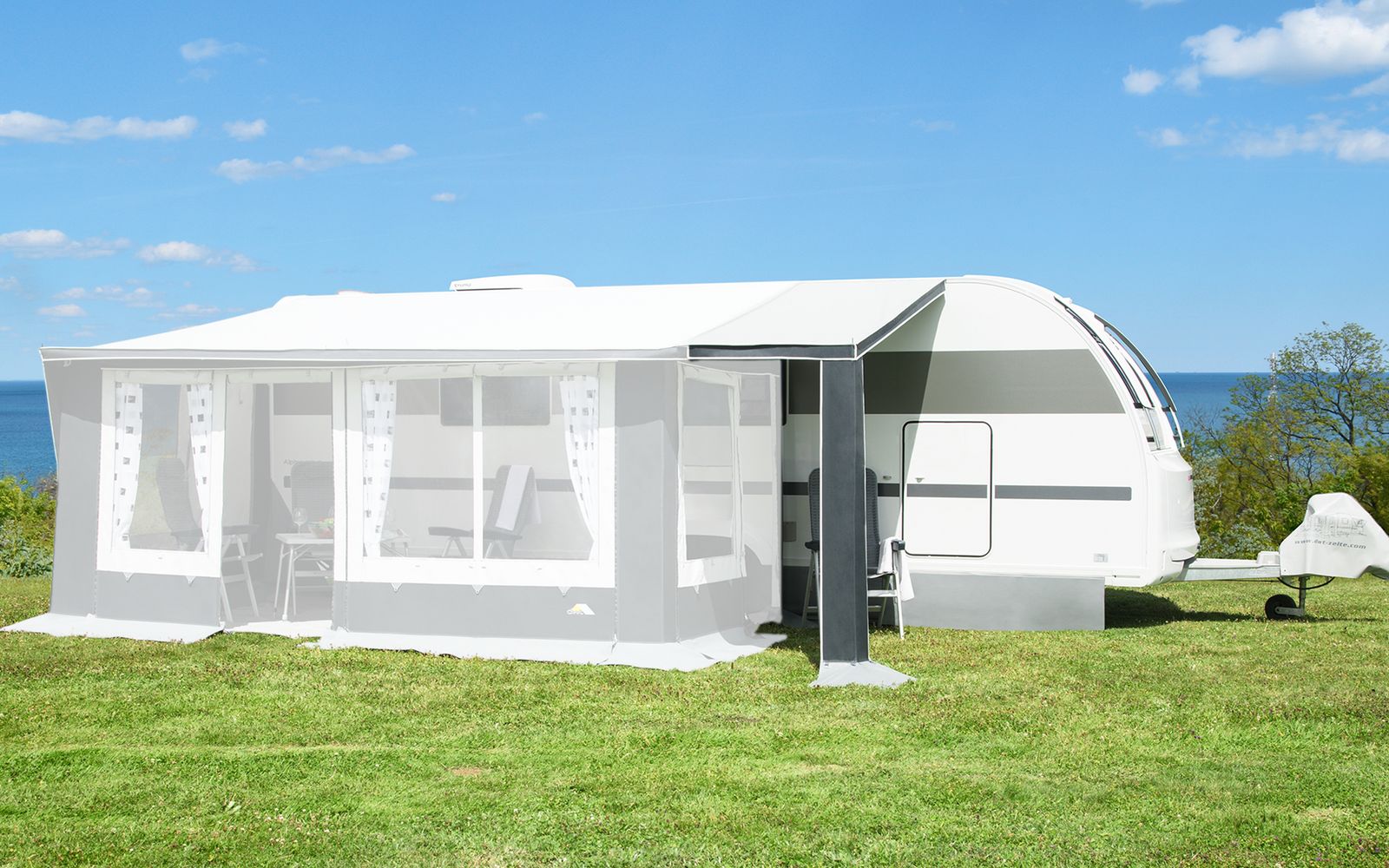 Weißer Wohnwagen mit angebautem Vorzelt auf einer Wiese, blauer Himmel mit wenigen Wolken, Meer und Bäume im Hintergrund