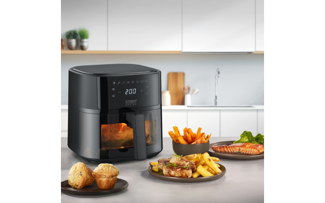 Schwarze Heißluftfritteuse mit digitaler Anzeige "200" auf einer Küchenarbeitsplatte; Lebensmittel im Sichtfenster. Auf der Arbeitsplatte: zwei Muffins auf einem Teller, ein Teller mit Steak, Pommes und Gemüse, eine Schale Pommes und ein Teller mit L