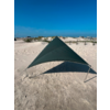 Dunkelgrünes, dreieckiges Tuch auf Metallstangen auf sandigem Strand, Schatten darunter, Dünen mit Gras und Gebäude im Hintergrund, blauer Himmel