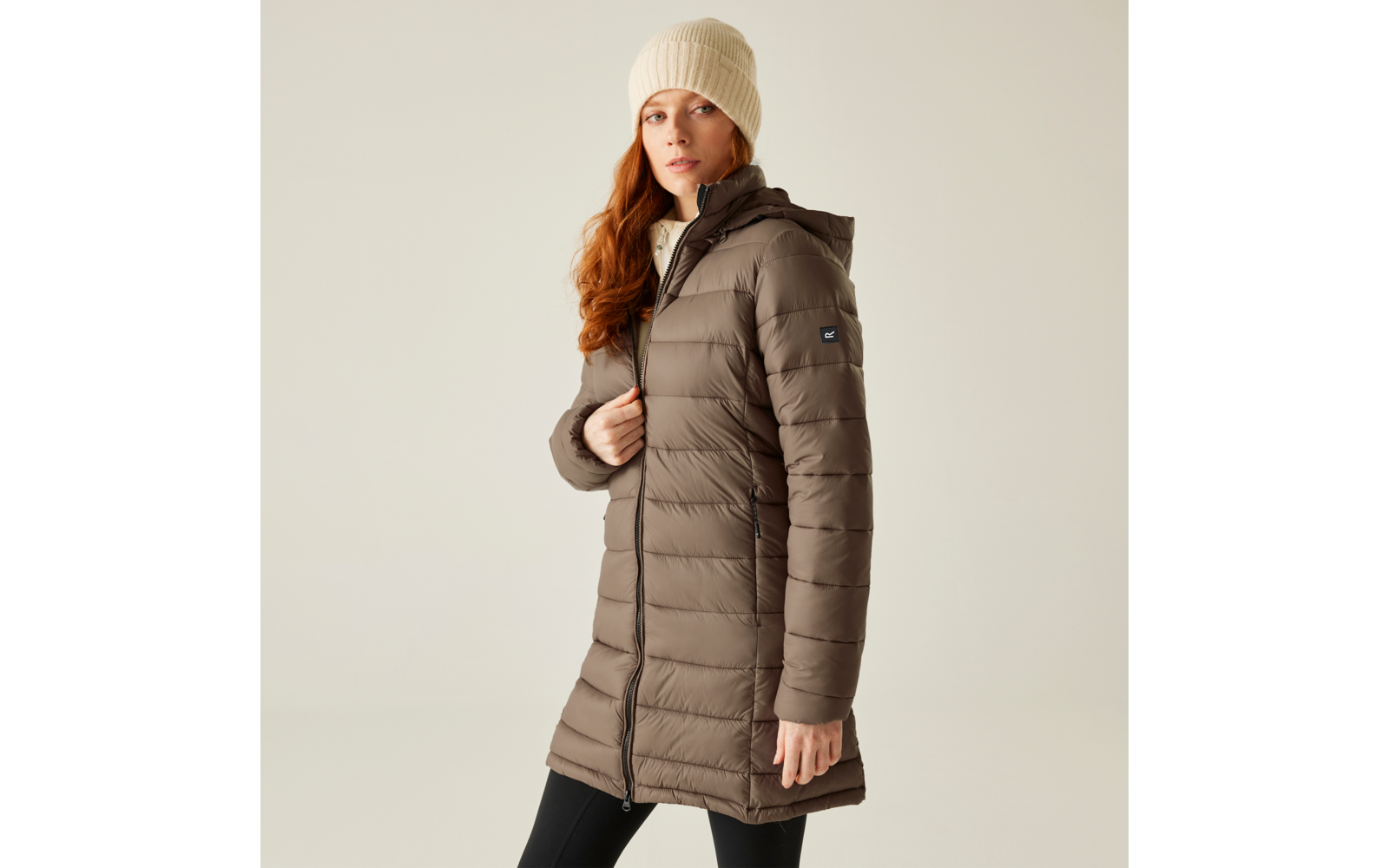 Frau mit rotem Haar trägt eine beige Mütze und eine lange braune Steppjacke
