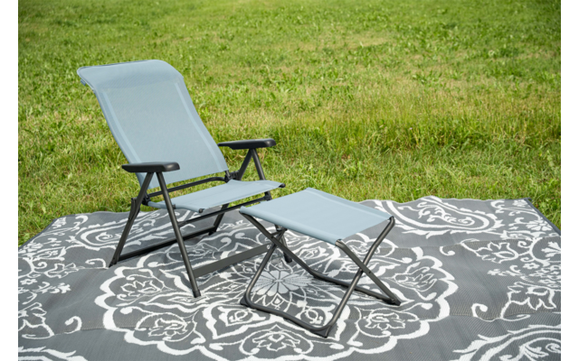 Fauteuil pliant bleu clair avec armature métallique noire et repose-pieds assorti sur un tapis d'extérieur gris à motif floral blanc, herbe en arrière-plan