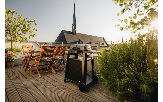 Dachterrasse mit Holzdeck, Holz-Gartenmöbel (Tisch, Klappstühle), Grill, Topfpflanzen; im Hintergrund Gebäudedach und Kirchturm, Sonnenschein von rechts