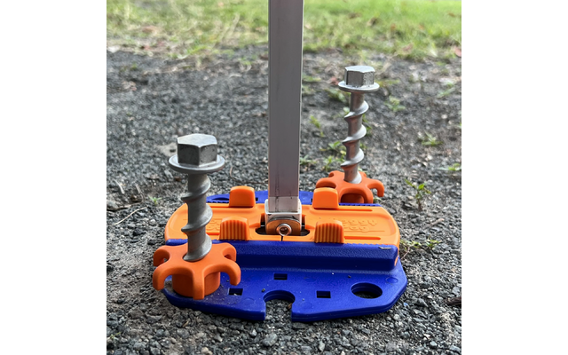 Blaue und orange Kunststoffplatte auf Schotter/Asphalt mit zwei großen, teilweise eingedrehten Metallschrauben mit Sechskantköpfen und einer senkrechten Metallstange, im Hintergrund Gras