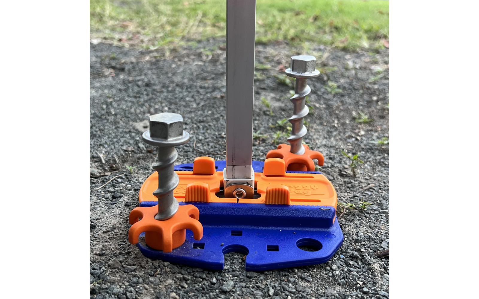 Blaue und orange Kunststoffplatte auf Schotter/Asphalt mit zwei großen, teilweise eingedrehten Metallschrauben mit Sechskantköpfen und einer senkrechten Metallstange, im Hintergrund Gras