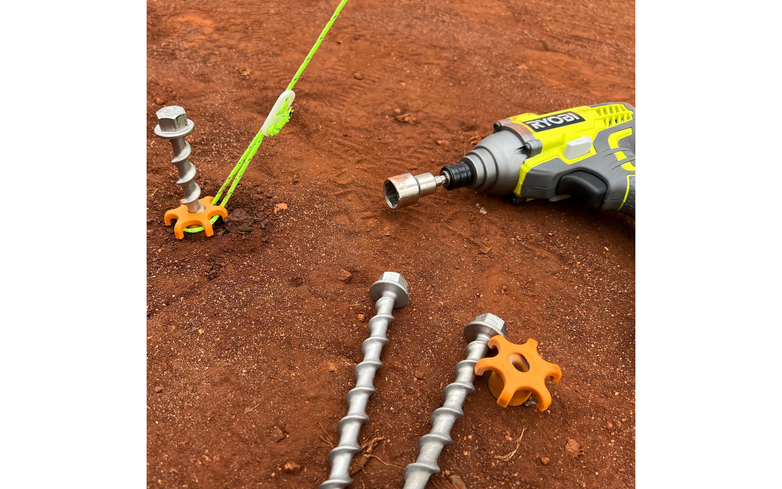 Gelber kabelloser Schrauber mit Steckaufsatz auf rotem Boden; drei spiralförmige Metallstangen; zwei orangefarbene sternförmige Kunststoffteile; neongrünes Seil, das an einer Stange befestigt ist