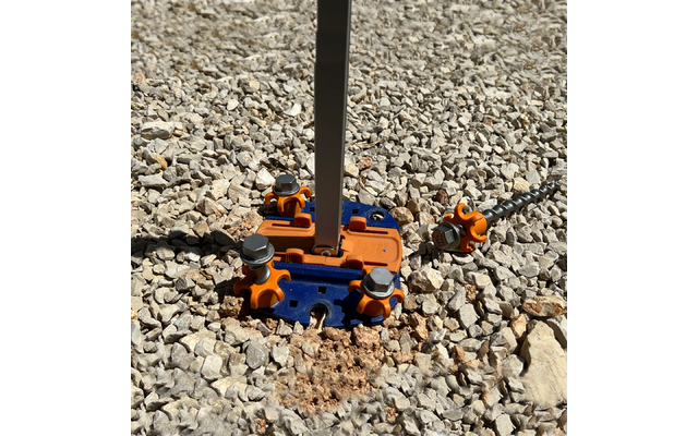 Senkrechte Metallstange in einem blauen und orangefarbenen Bauteil mit sichtbaren Sechskantmuttern auf kiesigem Schotter; daneben liegt ein weiteres blau-orange Bauteil mit Gewindestange