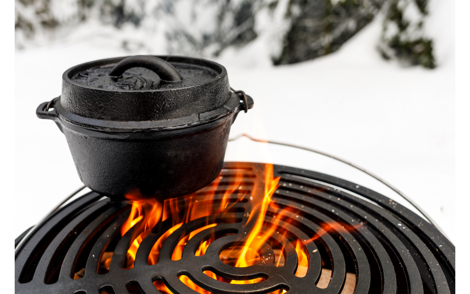 Schwarzer gusseiserner Topf mit Deckel auf rundem Grillrost, darunter Flammen; schneebedeckter Boden und unscharfe Bäume im Hintergrund