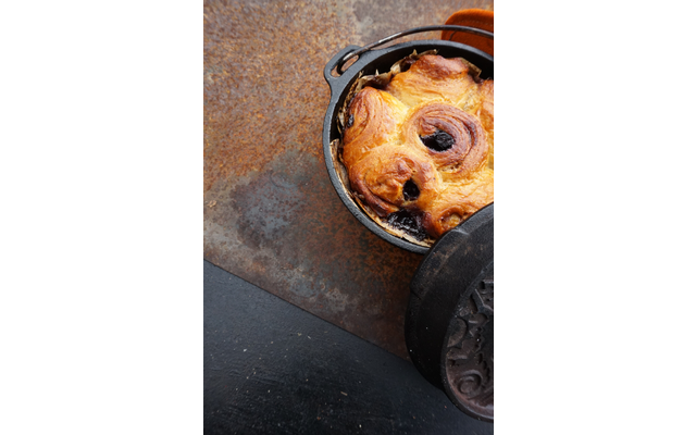 Runder schwarzer Gusseisentopf mit goldbraunem Gebäck mit dunkler Füllung, Deckel liegt teilweise darauf, auf rostiger Metallfläche und schwarzer Schieferplatte