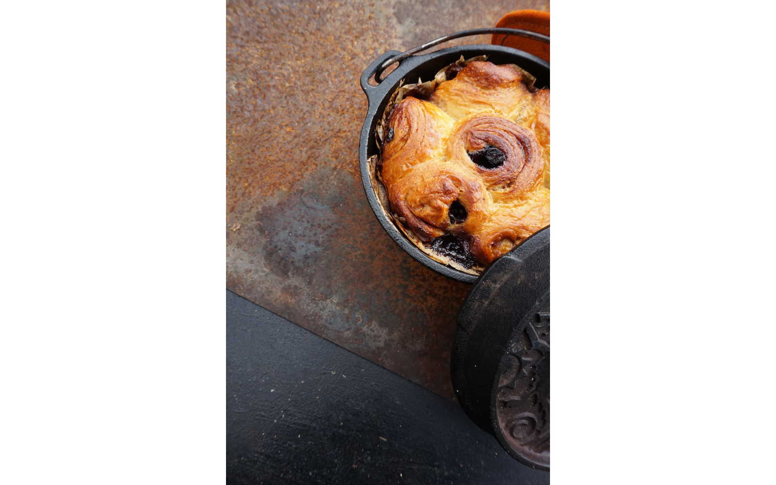 Runder schwarzer Gusseisentopf mit goldbraunem Gebäck mit dunkler Füllung, Deckel liegt teilweise darauf, auf rostiger Metallfläche und schwarzer Schieferplatte