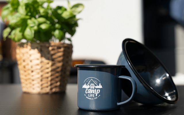 Tasse émaillée bleue avec l'inscription camp life et un bol émaillé noir incliné sur une table, à gauche un panier tressé avec une plante verte, arrière-plan flou