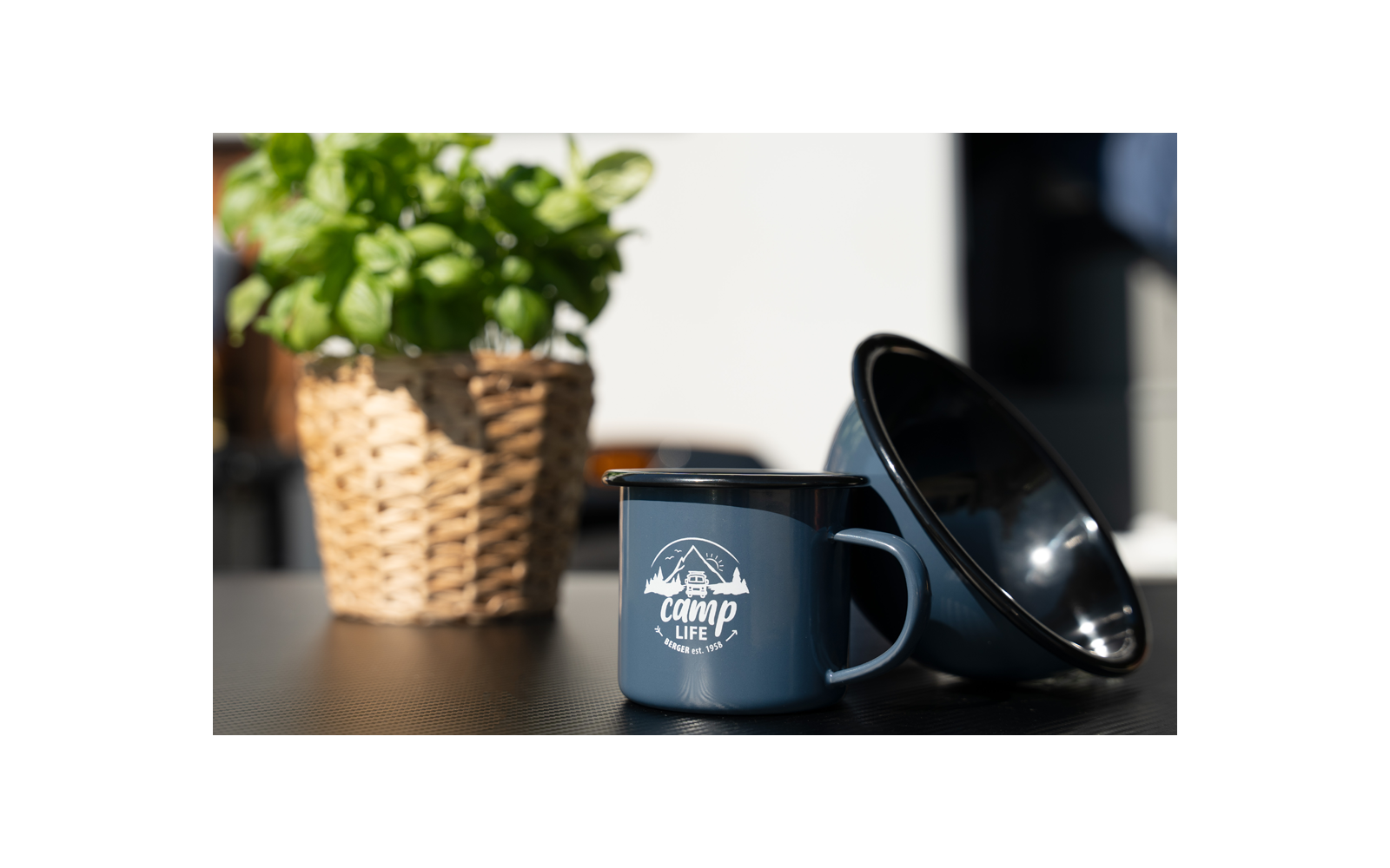 Tasse émaillée bleue avec l'inscription camp life et un bol émaillé noir incliné sur une table, à gauche un panier tressé avec une plante verte, arrière-plan flou