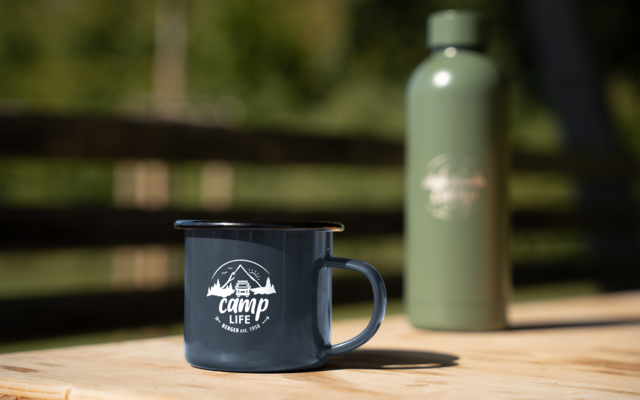 Blaue Emaille-Tasse mit Aufdruck "camp life" auf Holz­tisch, grüner Thermosbehälter im Hintergrund, unscharfer Zaun und Bäume