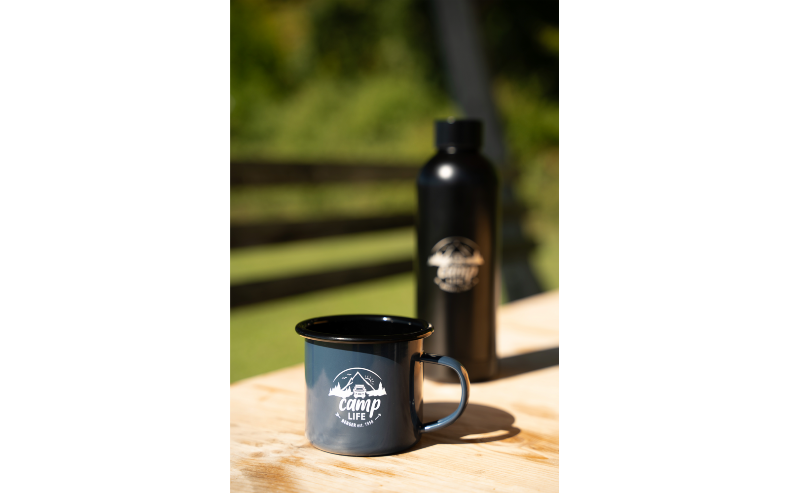 Dunkelblaue Emaille-Tasse mit weißer Aufschrift "camp life" und Berg-Logo auf einem Holztisch; dahinter eine schwarze Trinkflasche, verschwommene grüne Vegetation und horizontale Holzlatten
