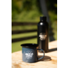 Dunkelblaue Emaille-Tasse mit weißer Aufschrift "camp life" und Berg-Logo auf einem Holztisch; dahinter eine schwarze Trinkflasche, verschwommene grüne Vegetation und horizontale Holzlatten
