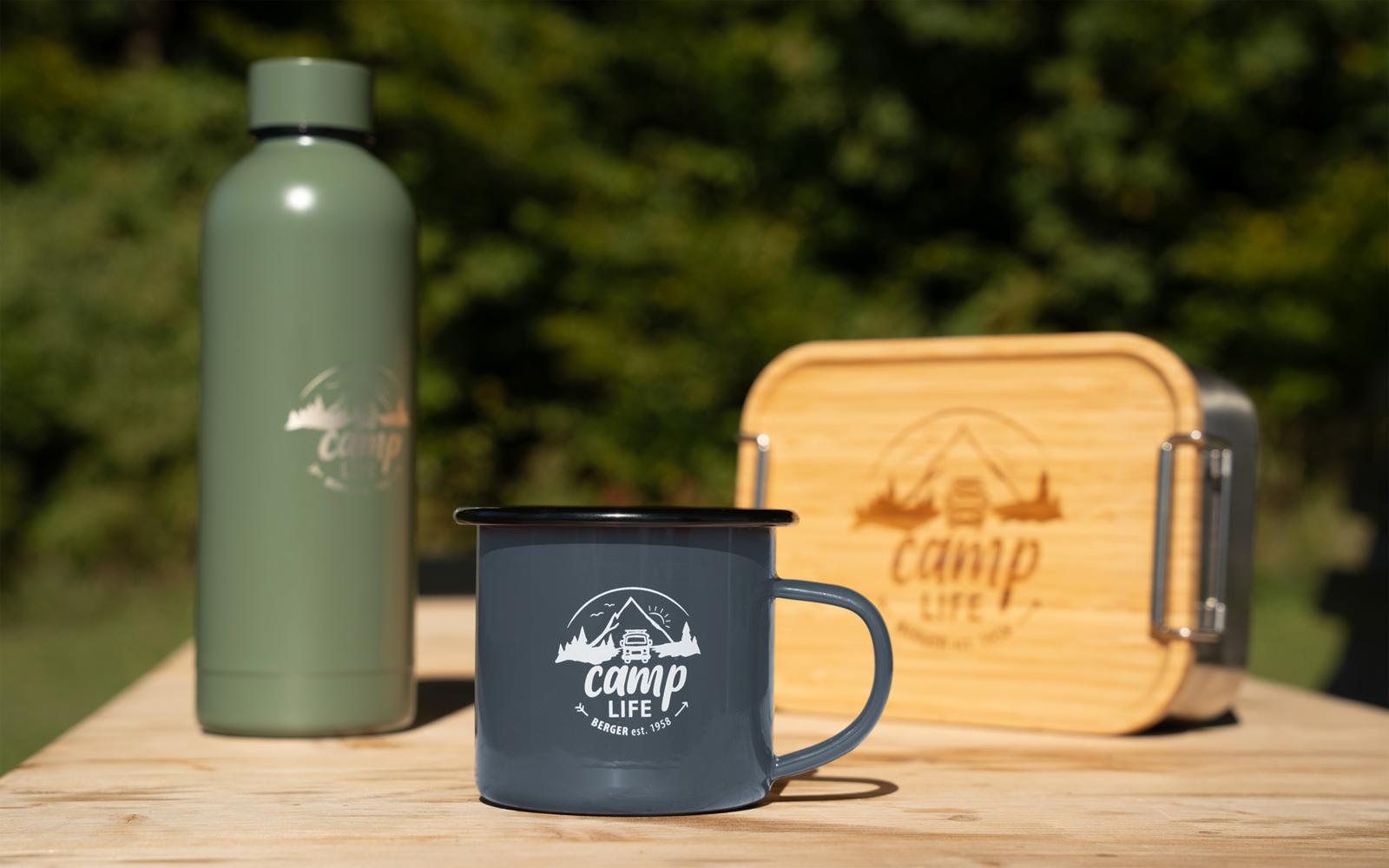 Graue Emaille-Tasse mit 'camp life'-Logo auf einem Holztisch, links eine grüne Thermosflasche, rechts eine rechteckige Holzbox mit Logo, unscharfer grüner Hintergrund mit Bäumen