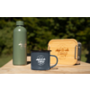 Graue Emaille-Tasse mit 'camp life'-Logo auf einem Holztisch, links eine grüne Thermosflasche, rechts eine rechteckige Holzbox mit Logo, unscharfer grüner Hintergrund mit Bäumen