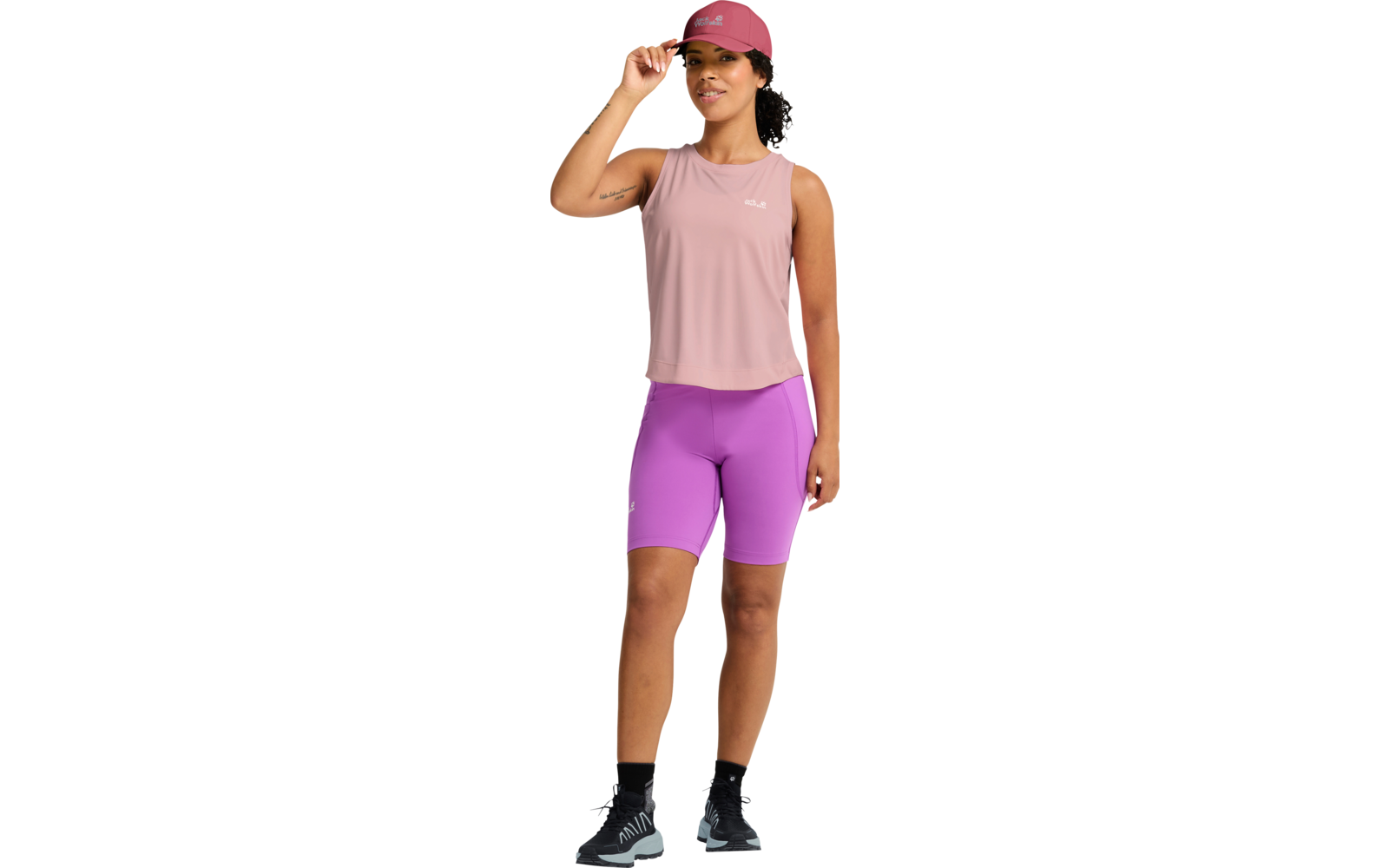 Person mit rosa Basecap, ärmellosem rosa Oberteil, hellvioletten Radshorts, schwarzen Socken und grauen Sportschuhen; stehend, eine Hand an der Cap-Krempe