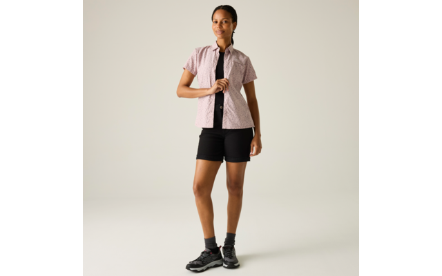 Frau steht frontal, trägt kurzärmeliges hellrosa gemustertes Hemd, schwarzes Top, schwarze Shorts, schwarze Socken und schwarze Sneaker vor hellem Hintergrund
