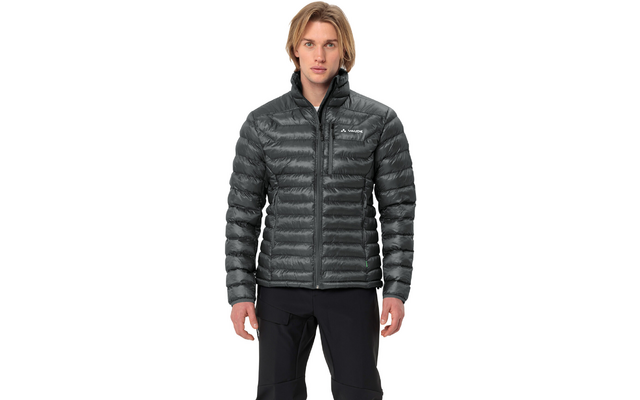 Person mit schulterlangen blonden Haaren trägt eine dunkle gesteppte Jacke mit Reißverschluss und sichtbarem Markenlogo, steht frontal vor weißem Hintergrund und schaut in die Kamera