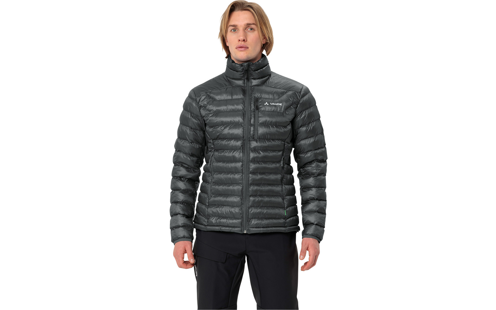 Mann mit schulterlangem blondem Haar, frontal, trägt dunkelgraue gesteppte Jacke mit hohem Kragen, Reißverschluss und Brustlogo; schwarze Hose; weißer Hintergrund
