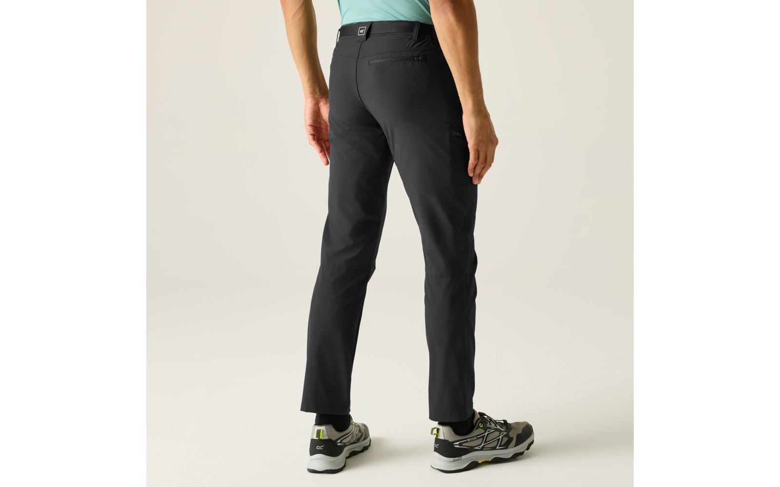 Rückenansicht einer stehenden Person, sichtbar von Taille bis Füßen, trägt schwarze Hose, hellgrünes Oberteil, graue Sneaker mit grünen Akzenten, heller neutraler Hintergrund