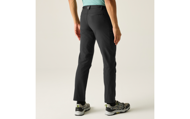 Rückenansicht einer stehenden Person, sichtbar von Taille bis Füßen, trägt schwarze Hose, hellgrünes Oberteil, graue Sneaker mit grünen Akzenten, heller neutraler Hintergrund