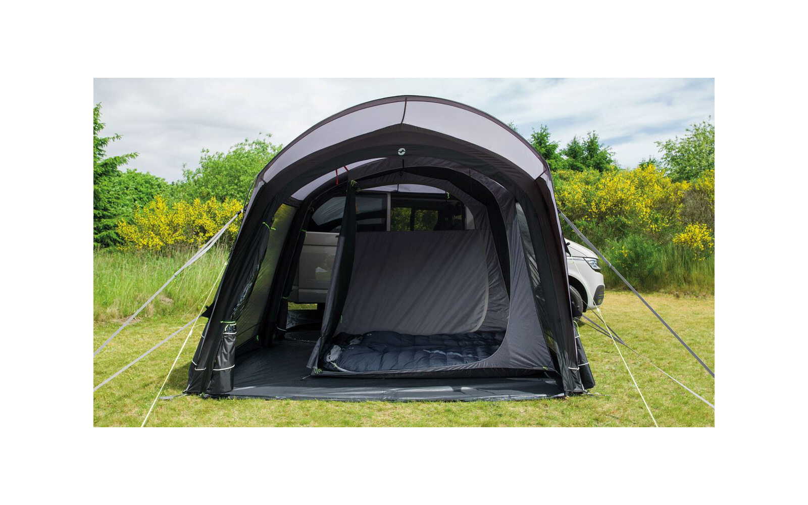 Aufgeschlagener dunkelgrauer aufblasbarer Campingvorbau mit offenem Eingang und einem Schlafsack auf dem Boden; rechts teilweise sichtbares weißes Auto; Grasfläche, gelbe blühende Büsche und Bäume im Hintergrund, bewölkter Himmel