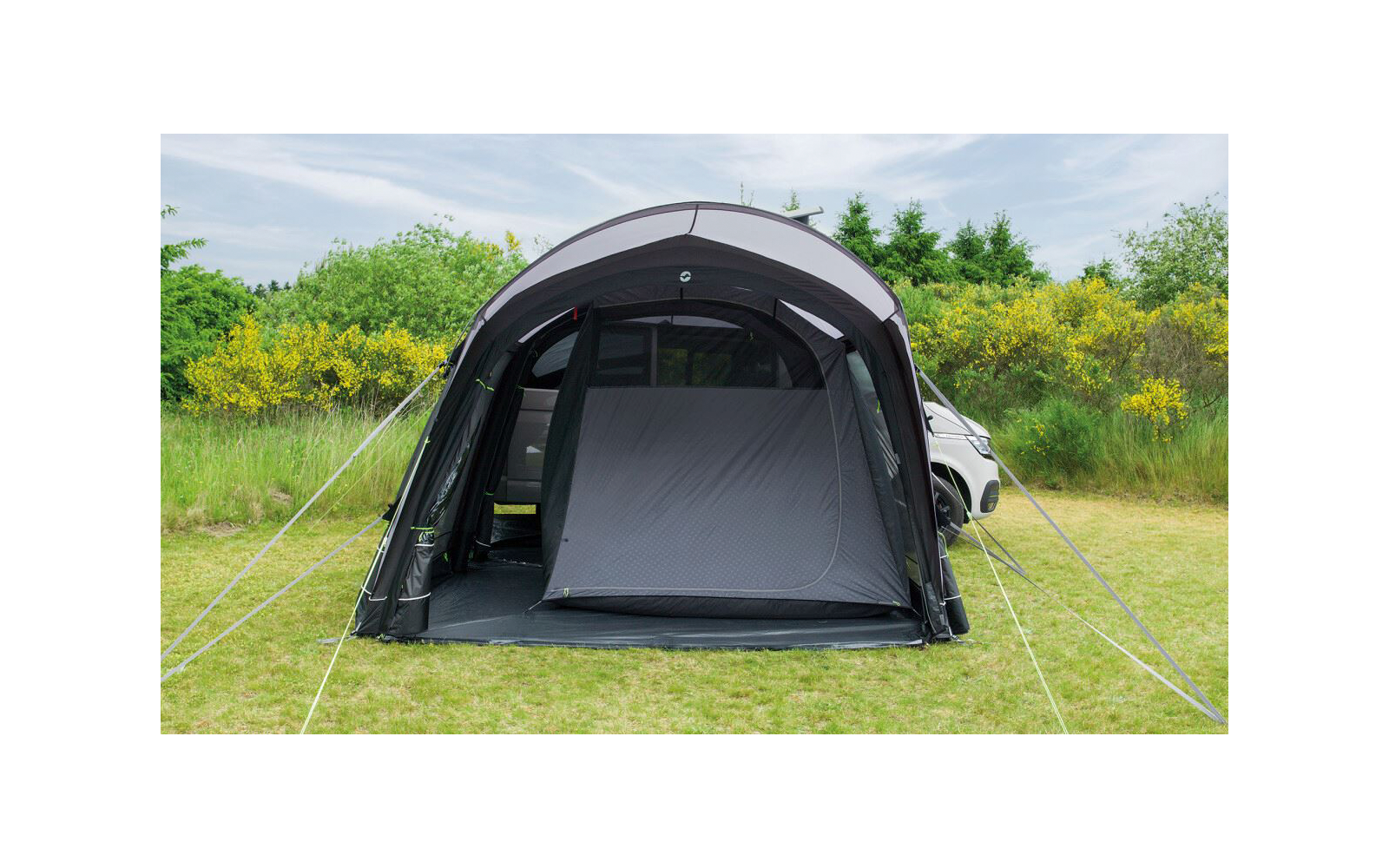 Dunkles aufblasbares Vorzelt neben einem weißen Auto auf einer Wiese; Vorderseite mit geschlossener Innenkabine, seitlichen Öffnungen, Abspannleinen und gelb-grünem Gebüsch im Hintergrund